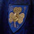 Close-up of the embroidered logo on the St. Patrick Days 2025 Jersey featuring a blue shield with a golden shamrock and decorative elements.