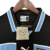 Camisa retrô da Lazio de 1998, da marca Puma, com detalhes em preto e azul claro, pendurada em um cabide.