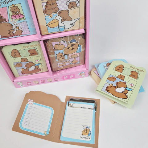 Bloquinho de papel com estampa de capivara e clipe metálico, exibindo várias capas coloridas e um bloco aberto com espaço para anotações.