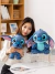 Mulher segurando dois brinquedos de pelúcia do Stitch, um pequeno e um maior, em um ambiente aconchegante.