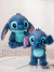 Dois bonecos de pelúcia do personagem Stitch, um maior e um menor, com pelagem azul e detalhes em verde, sentados sobre uma grade de metal.