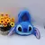 Bolsa mochila pelúcia do Stitch com orelhas e olhos grandes, ao lado de um vaso com flores amarelas.