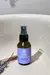 Frasco de vidro âmbar do Desodorante Natural Spray Levide de 60ml, com borrifador preto e rótulo roxo. O frasco está posicionado em um ambiente úmido, com gotas de água visíveis em uma superfície de louça clara e galhos de lavanda seca na lateral, sugerin