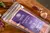 Sachê de 200ml do hidratante Argiloterapia Levidè com rótulo roxo sobre superfície de madeira, decorado com folhas e conchas