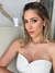 Mulher com cabelo liso e loiro, usando um vestido branco sem alças e colar dourado, posando em um ambiente moderno.