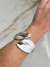 Bracelete prateado com design moderno, mostrando detalhes de folhas, posicionado no pulso de uma pessoa.