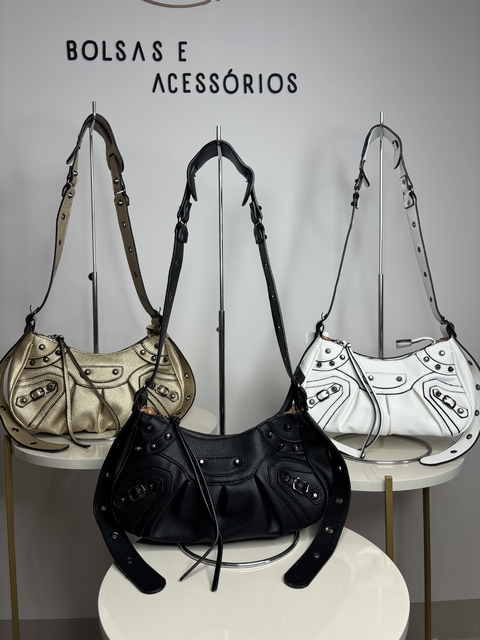 Três bolsas em exibição: uma dourada, uma preta e uma branca, todas com detalhes em metal e alças ajustáveis.
