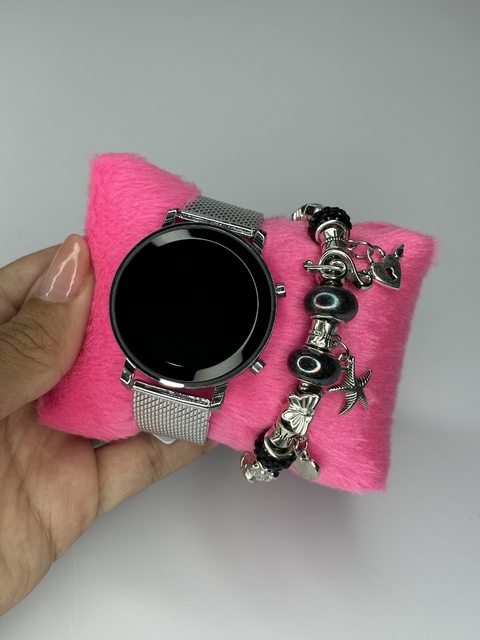 Relógio digital redondo com pulseira prata, apresentado sobre um suporte rosa de pelúcia.