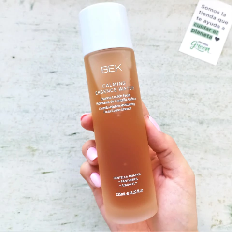 Botella de esencia facial hidratante y antioxidante de BEK, con tapa blanca y líquido de color ámbar, sostenida en una mano.