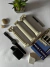 Tres columnas decorativas clásicas en una mesa con un reloj negro, un frasco de perfume, un libro sobre Grecia y una planta en maceta.