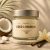 Vela aromática em pote de vidro com tampa metálica, fragrância coco e baunilha, pesando 150g, com coco cortado e flor de baunilha ao lado.