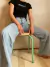 Una persona sentada sobre un banquito de diseño moderno con patas verdes y asiento de madera, vistiendo una camiseta negra y pantalones de mezclilla claros.