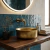 Bacha de baño dorada brillante sobre encimera de madera con grifería de pared y fondo de azulejos azules