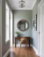 A circular wooden mirror in a hallway with a wooden table, a vase, and a chair.