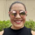 Mulher sorrindo usando óculos de sol rosa com lente rosa, em um ambiente externo com vegetação ao fundo.