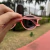 Óculos Baixa Pace Round Two na cor rosa, com lentes rosa, sendo segurados por uma mão em um ambiente externo.