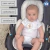 Imagem de um bebê em um carrinho, usando uma capa anatômica de tecido 100% algodão. O bebê está com um macacão branco e tem uma expressão neutra.