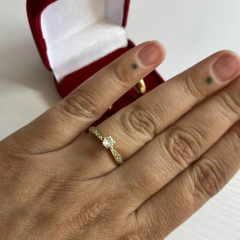 Anel solitário dourado cravejado com pedra central, usado no dedo da mão esquerda, com caixa vermelha de veludo ao fundo