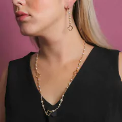 Modelo usando um conjunto de semijoias com pérolas e trevos vazados, incluindo um colar e brincos, em fundo rosa.