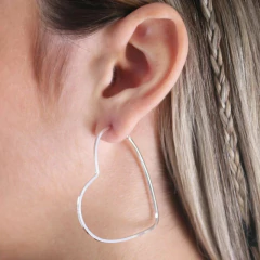 Brinco argola em formato de coração, feito em prata 925, sendo usado na orelha de uma mulher com cabelo liso.