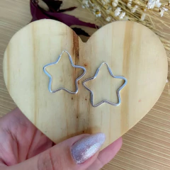 Brinco argola em forma de estrela, feito em prata 925, apresentado sobre um suporte de madeira em formato de coração.