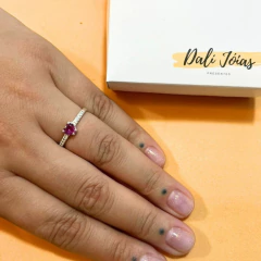 Anel solitário cravejado com coração pink em uma mão sobre fundo laranja, com embalagem ao fundo.