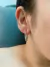 Close-up de uma orelha feminina com brinco em ródio branco em forma de argola aberta retangular. MW Semijoias e acessórios
