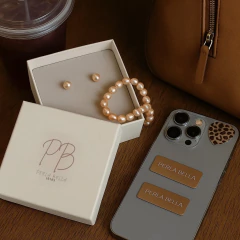 Conjunto de colar e brinco de pérola pêssego em uma caixa elegante, ao lado de um copo e um celular com adesivo de onça.