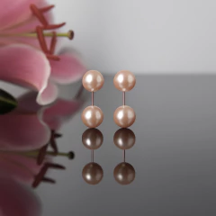 Brinco Gemini pêssego com duas pérolas redondas em tom pêssego, refletido em superfície espelhada com flor rosa desfocada ao fundo