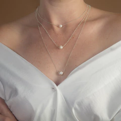 Colar delicado com três correntes finas em prata, cada uma com uma pérola branca em tamanhos variados, usado por uma pessoa com blusa branca decotada