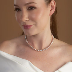 Mulher usando colar de pérolas da Coleção Lírios, modelo Colar Doce Marfim, com vestido branco e fundo marrom