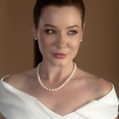 Mulher usando colar de pérolas brancas e brinco de pérola, com vestido branco e fundo marrom