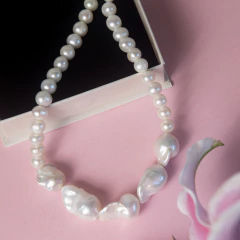 Colar de pérolas brancas com design barroco, exibido sobre fundo rosa com caixa preta e flor