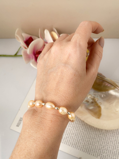 Pulseira Ilha Bela em uma mão, com pérolas e detalhes prateados, em um fundo decorativo com orquídeas.