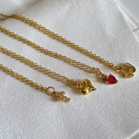 Collares dorados con dijes en forma de cruz, corazón y una joya roja, sobre un fondo blanco.