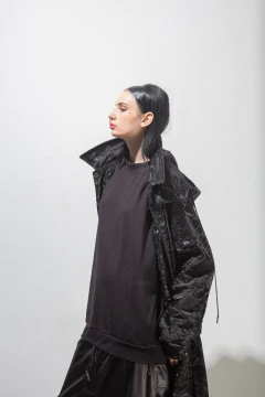A persona con cabello largo y oscuro lleva un abrigo largo negro de textura brillante, camiseta negra y pantalones oscuros, posando sobre un fondo blanco.