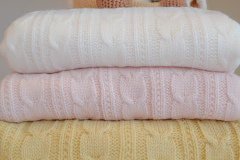 Três cobertores de bebê grossos de tricô com padrão de trança empilhados nas cores branco, rosa claro e amarelo claro