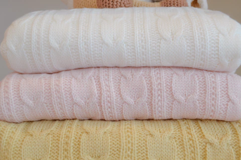 Três cobertores de bebê grossos de tricô com padrão de trança empilhados nas cores branco, rosa claro e amarelo claro