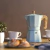Cafetera italiana verde Brinox Verona de 6 tazas y 300 ml sobre una mesa, con tazas de café y medialunas al fondo