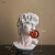 Escultura decorativa de Michelangelo com uma bola de chiclete vermelha na boca, destacando detalhes clássicos e contemporâneos.