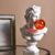 Escultura decorativa de um busto grego com uma bola de chiclete laranja na boca, ao lado de uma moldura de foto.
