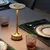 Luminária de mesa sem fio na cor dourada, iluminando uma mesa com copos e pratos elegantes.