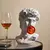 Escultura decorativa em forma de busto, inspirada em Michelangelo, com uma bola de chiclete vermelha na boca, ao lado de um copo de bebida.