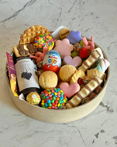Cesta de café da manhã chamada Cesta Magia com biscoitos decorados, suco de uva, ovo Kinder, doces coloridos, pães de queijo e chocolates variados em embalagem redonda