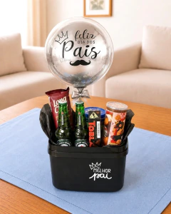 Cesta de Dia dos Pais com balão prata escrito "Feliz Dia dos Pais", duas garrafas de cerveja Heineken, chocolates e snacks em uma cesta preta sobre uma mesa com toalha azul.
