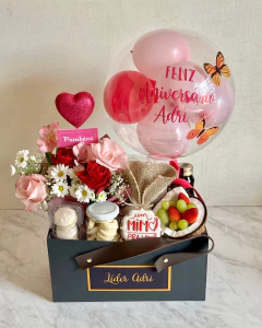 Box cartonada premium com arranjo de flores vermelhas, rosas e brancas, balão transparente com detalhes em rosa e vermelho, frutas frescas, doces e garrafa de bebida em embalagem preta com alça de couro
