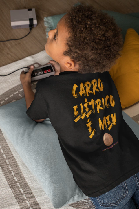 Criança deitada brincando com controle de videogame vestindo camiseta infantil preta com estampa amarela "Carro elétrico é meu"