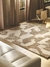 Alfombra Breeze con diseño abstracto en tonos beige y marrón, colocada en un ambiente moderno con un sofá y cortinas suaves.