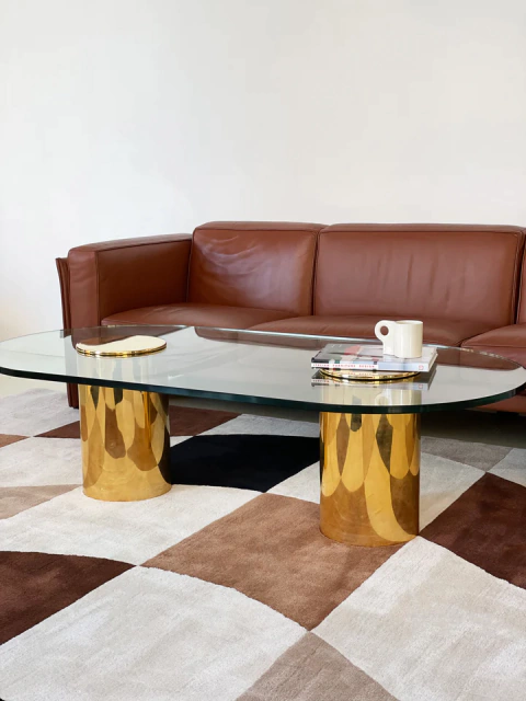 Alfombra Áurea con diseño geométrico en tonos marrones y crema, junto a una mesa de vidrio y una silla de cuero marrón.