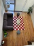 Vista aérea de una alfombra Damero de diseño a cuadros rojos y blancos, acompañada de un sofá gris y una mesa de vidrio en un ambiente moderno.